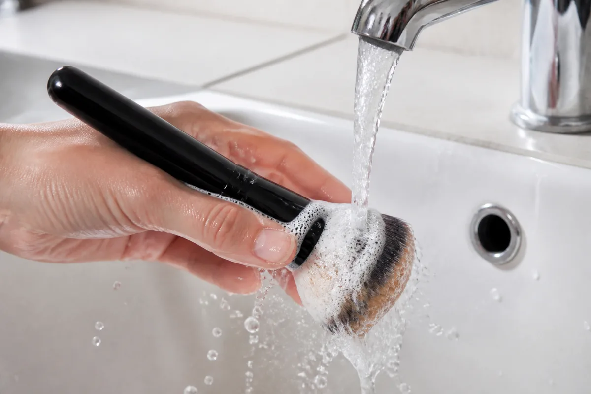 Pinceau à fond de teint rincé sous un filet d’eau tiède, mousse blanche visible sur les poils